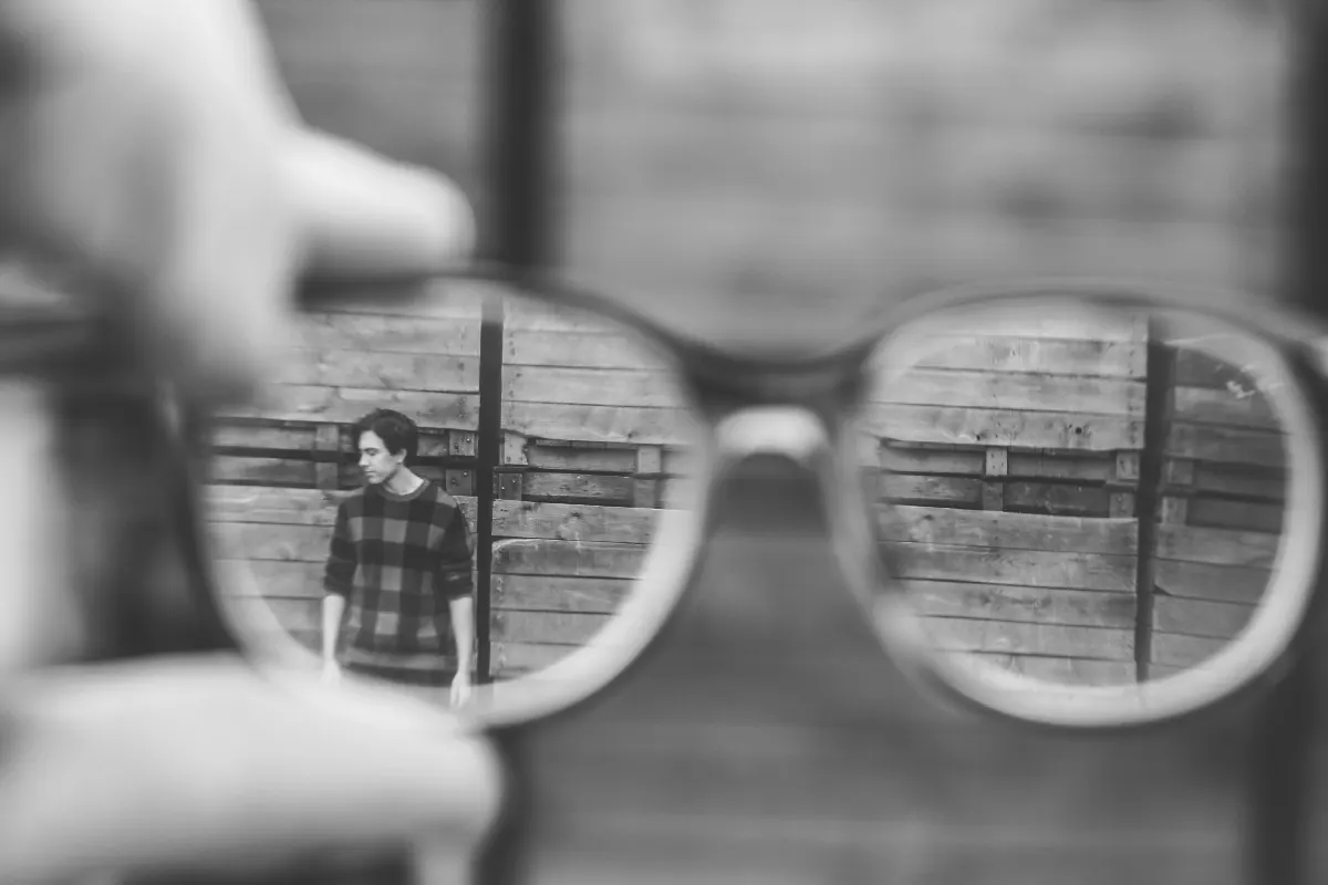 Photo by Joanna Nix on Unsplash. Image description: Black and white photo taken through a pair of glasses held up to the camera, of a man standing against a wall in the distance. The image is out of focus.